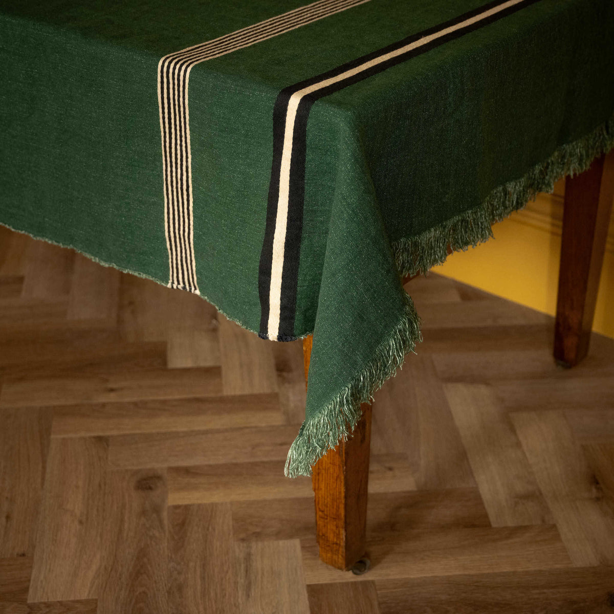 Libeco Belgian Linen 'Nell' Pièce di Linj. A dark green tablecloth with fringe and striped borders draped over a wooden table on a parquet wood floor.