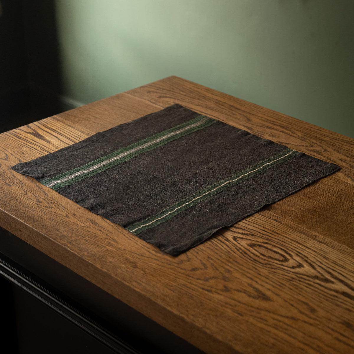 Libeco Belgian Linen 'Stella' Napkin. A faded black base with racing green, bone and charcoal stripes, displayed on a rustic wooden table.