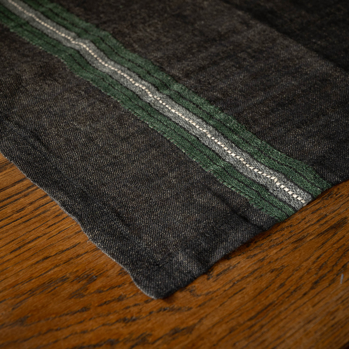 A close-up of Libeco Belgian Linen 'Stella' Napkin. A faded black base, featuring horizontal multi stripes of racing green, bone and charcoal grey, laid flat on a wooden surface.