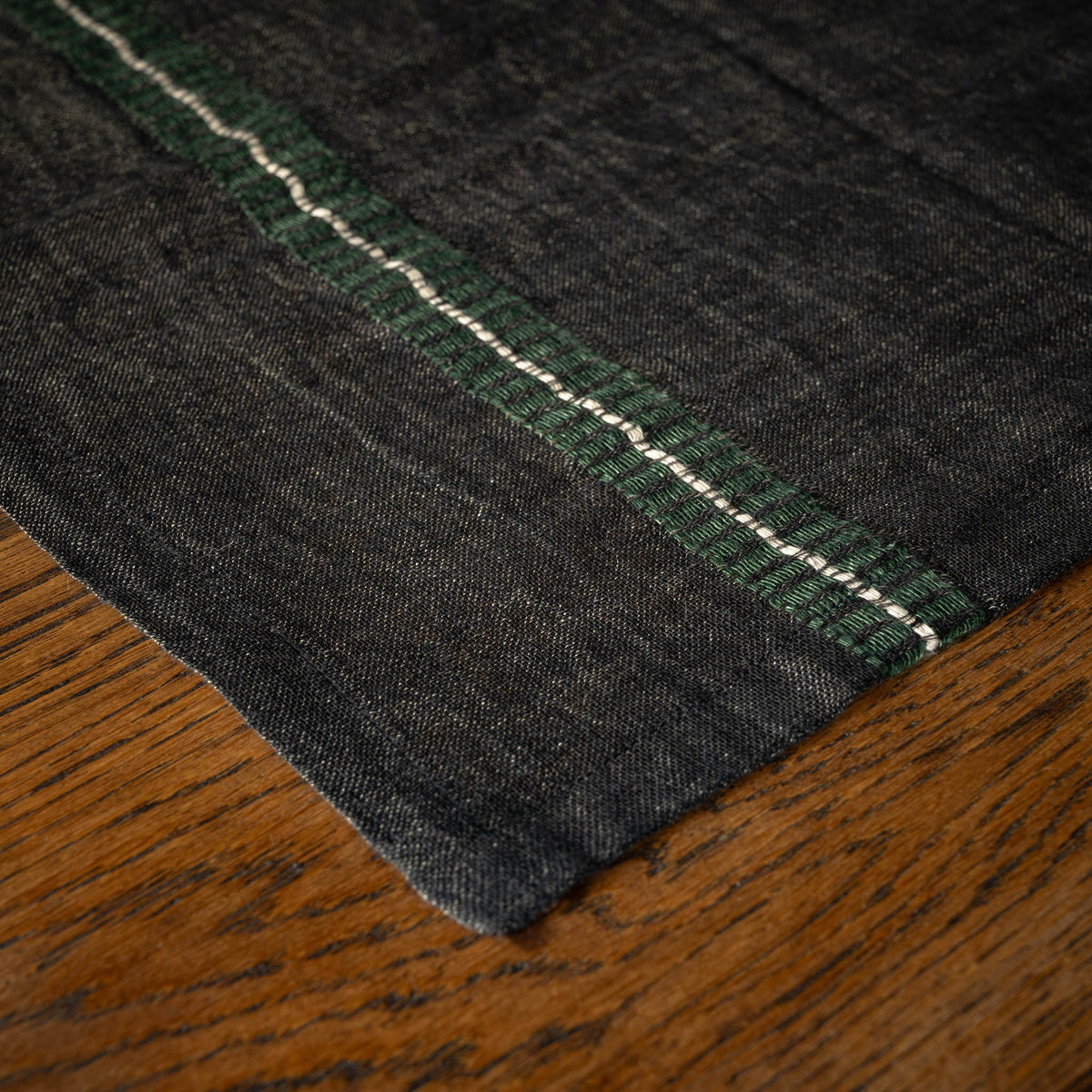 Close-up of Libeco Belgian Linen 'Stella' Napkin. A faded black base  with a racing green and bone stripe, resting on a brown wooden surface.