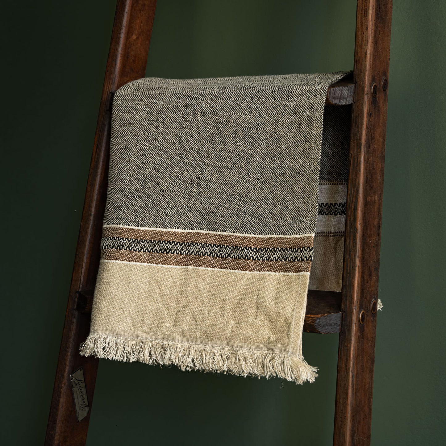 Libeco Linen hand towel draped over a dark wooden ladder against a dark green wall. The towel features a textured grey upper section, a cream lower section with fringe, and a decorative horizontal stripe pattern in the middle.