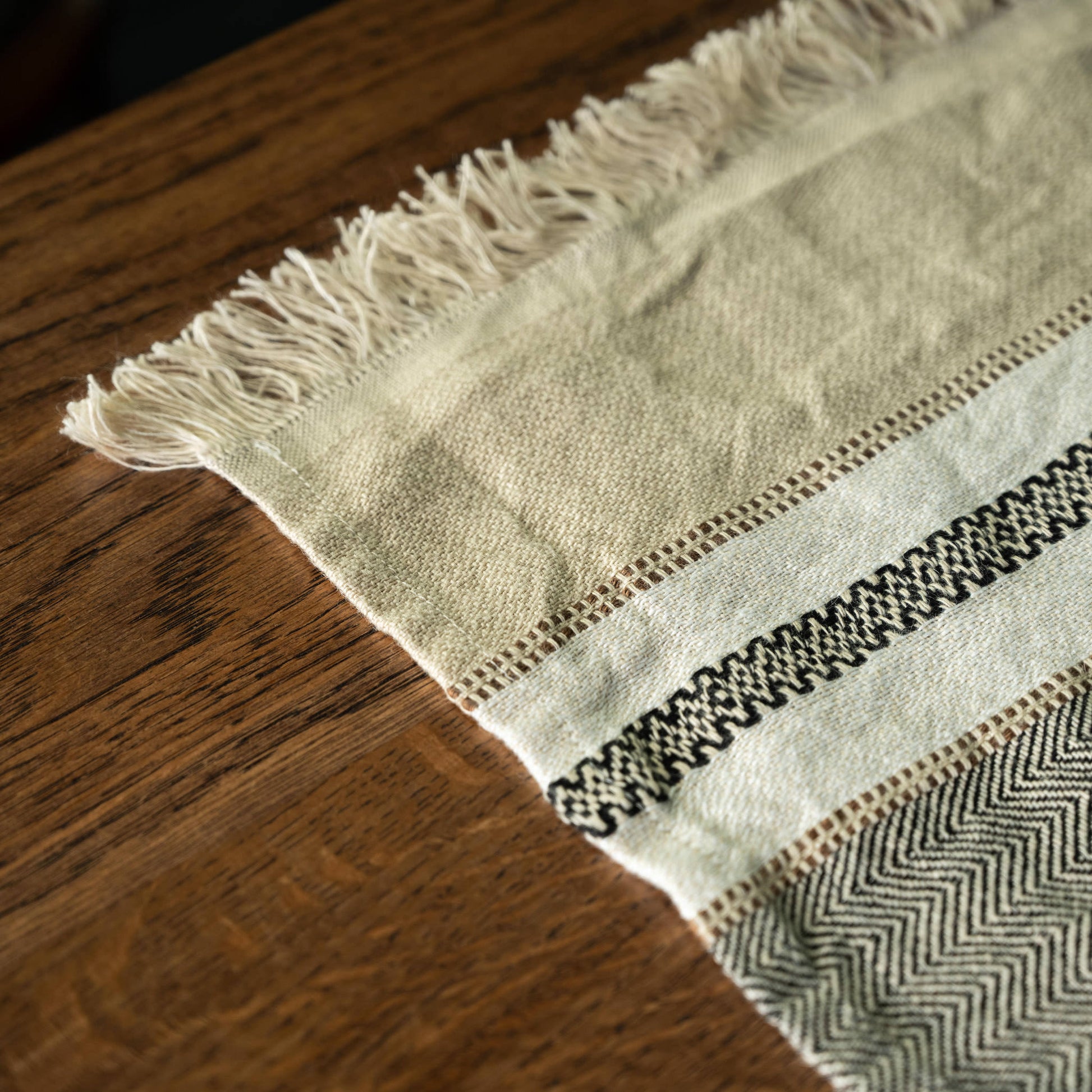 A close-up Libeco Linen guest towel laid diagonally across a dark brown wooden table. The towel has cream-coloured fringe on one end, a central cream band with a black geometric woven stripe, and a herringbone patterned section in black and off-white.