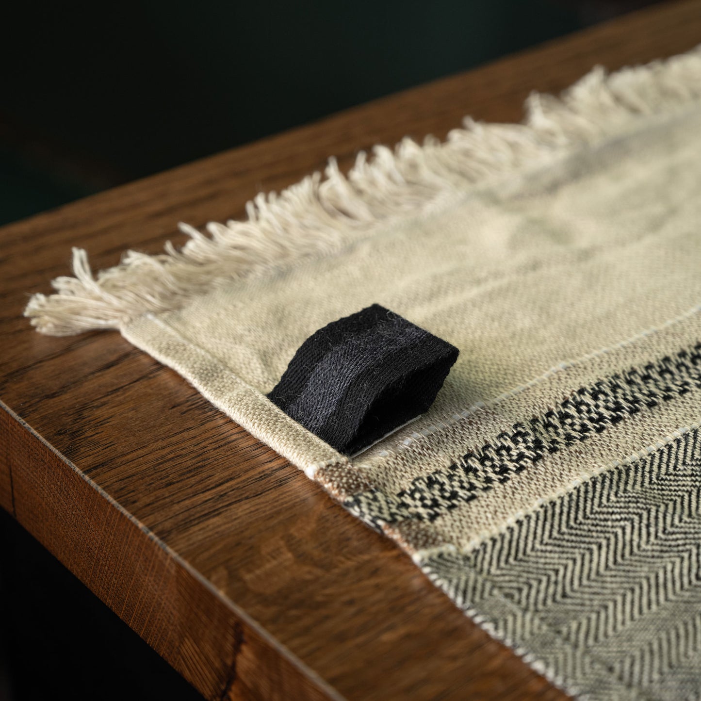 A close-up Libeco Linen guest towel  laid across a wooden table. The towel is off-white/cream with a black woven band, fringe along one edge, and a black fabric loop to hang it from.