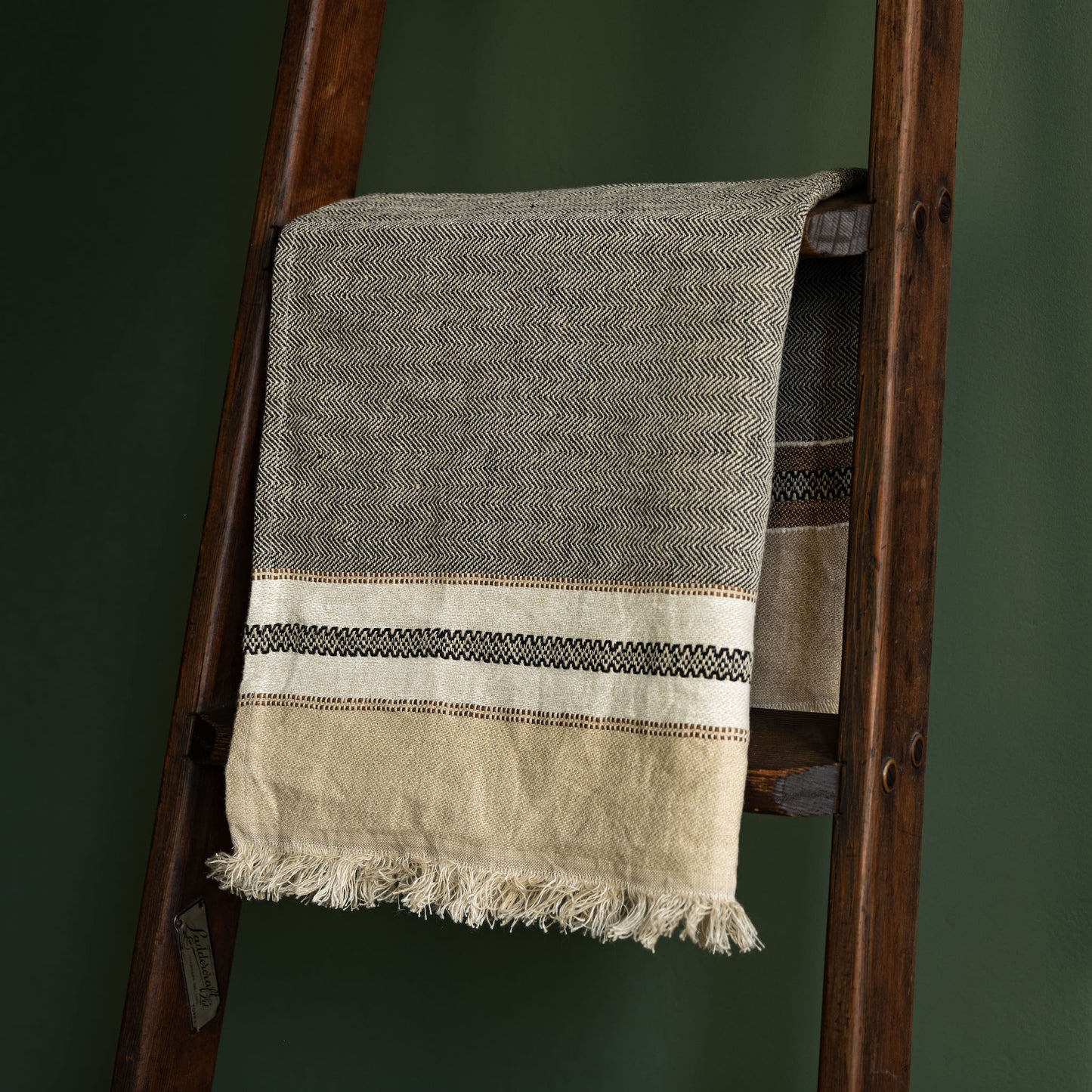 Libeco Linen hand towel in grey and beige with a striped border, draped over a dark wooden ladder against a dark green wall.