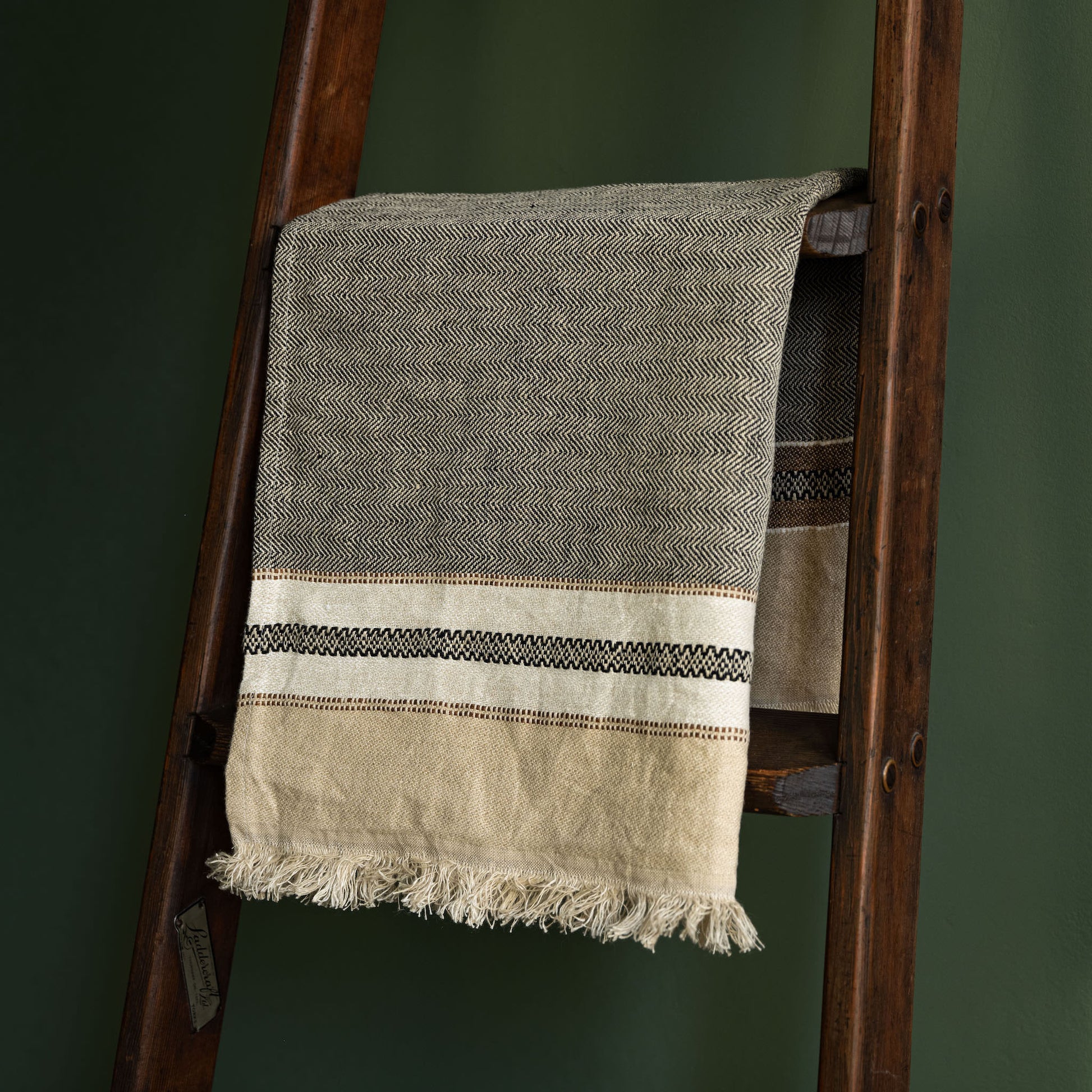 Libeco Linen hand towel in grey and beige with a striped border, draped over a dark wooden ladder against a dark green wall.