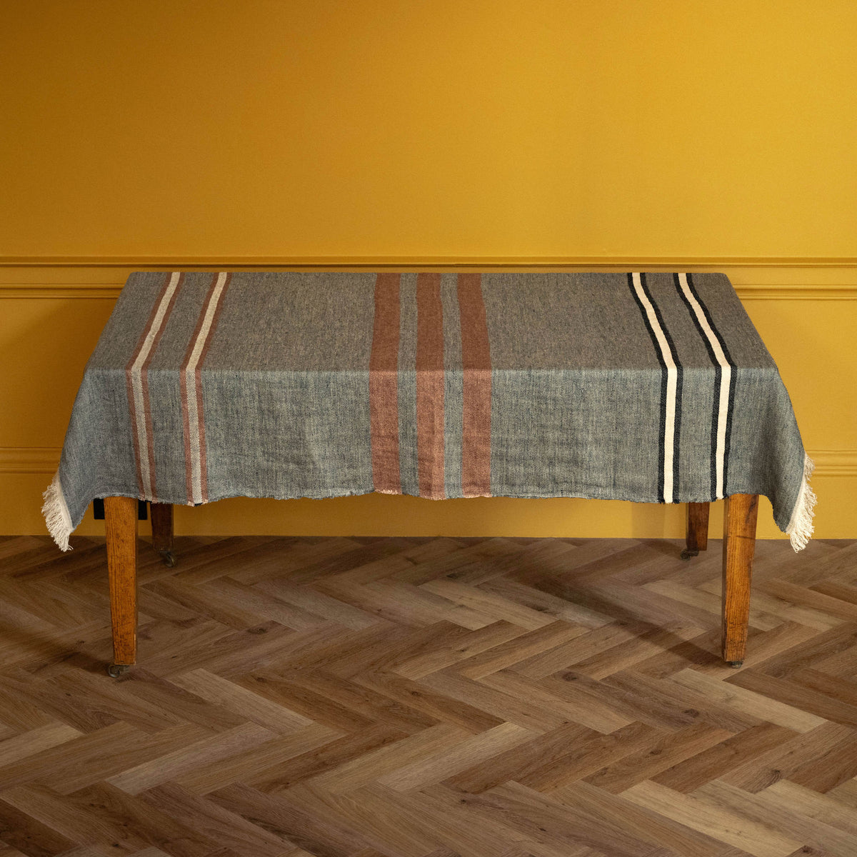 Libeco Belgian Linen Towel. A rectangular woven linen throw / tablecloth with fringe edges, featuring a heather grey base with vertical stripes in sienna-brown, black, and ecru, draped over a wooden table against a mustard yellow wall and herringbone wood floor.