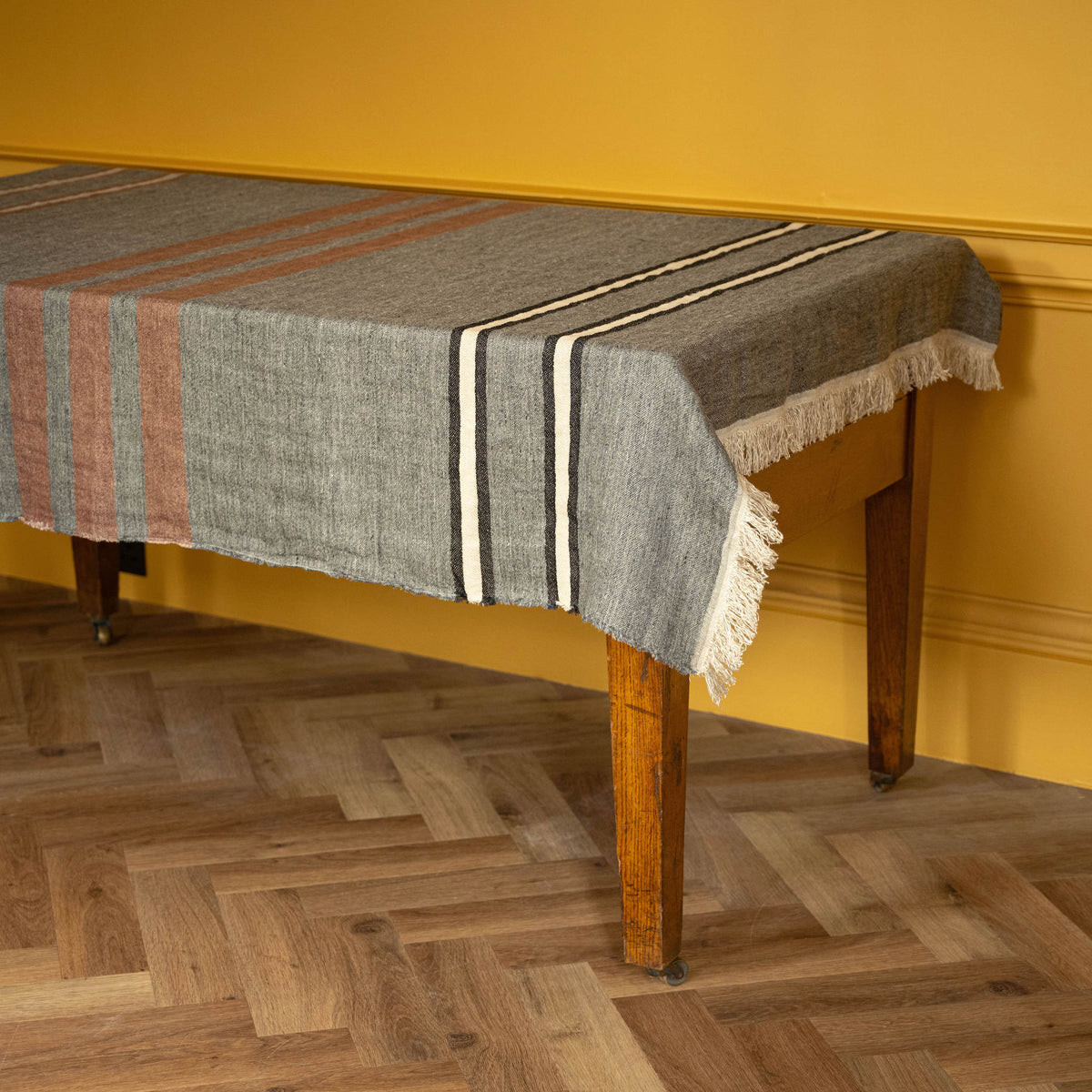 Libeco Linen Belgian Towel. A woven linen throw with fringe edge, featuring a grey base with sienna brown, ecru, and black stripes, draped over a wooden table against a mustard yellow wall and herringbone wood floor.