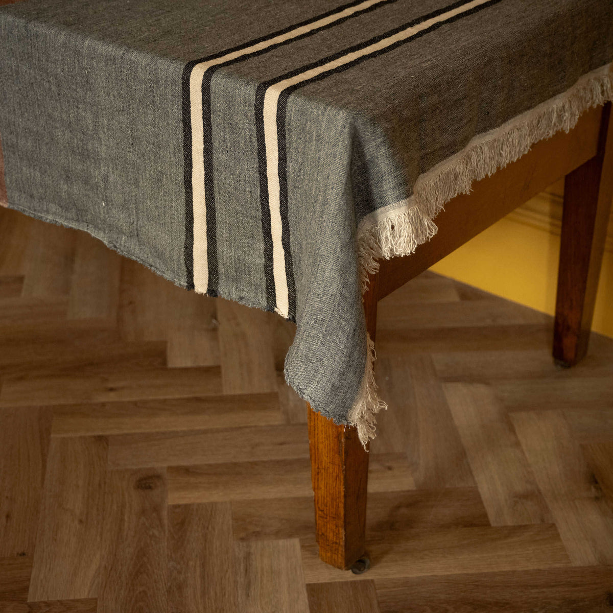 Libeco Linen Belgian Towel. A heater grey woven throw with ecru and black stripes and fringe edge draped over a wooden table on a herringbone wood floor.