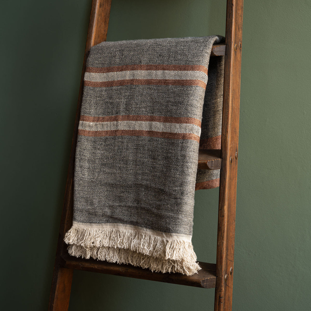 Libeco Linen Belgian Towel. A woven heather grey linen throw with horizontal stripes in sienna brown and cream, draped over a rustic wooden ladder against a dark green wall.
