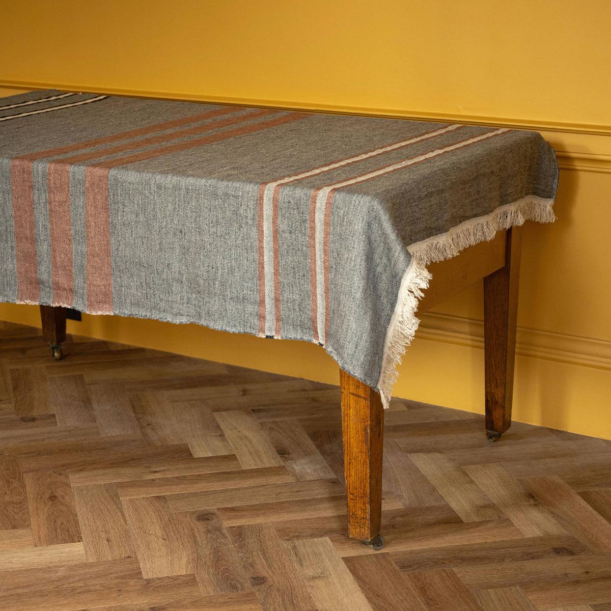 Libeco Linen Belgian Towel. Woven linen throw with fringe edge, heather grey base, sienna brown and ecru stripes, draped over a wooden table against a mustard yellow wall and herringbone wood floor.