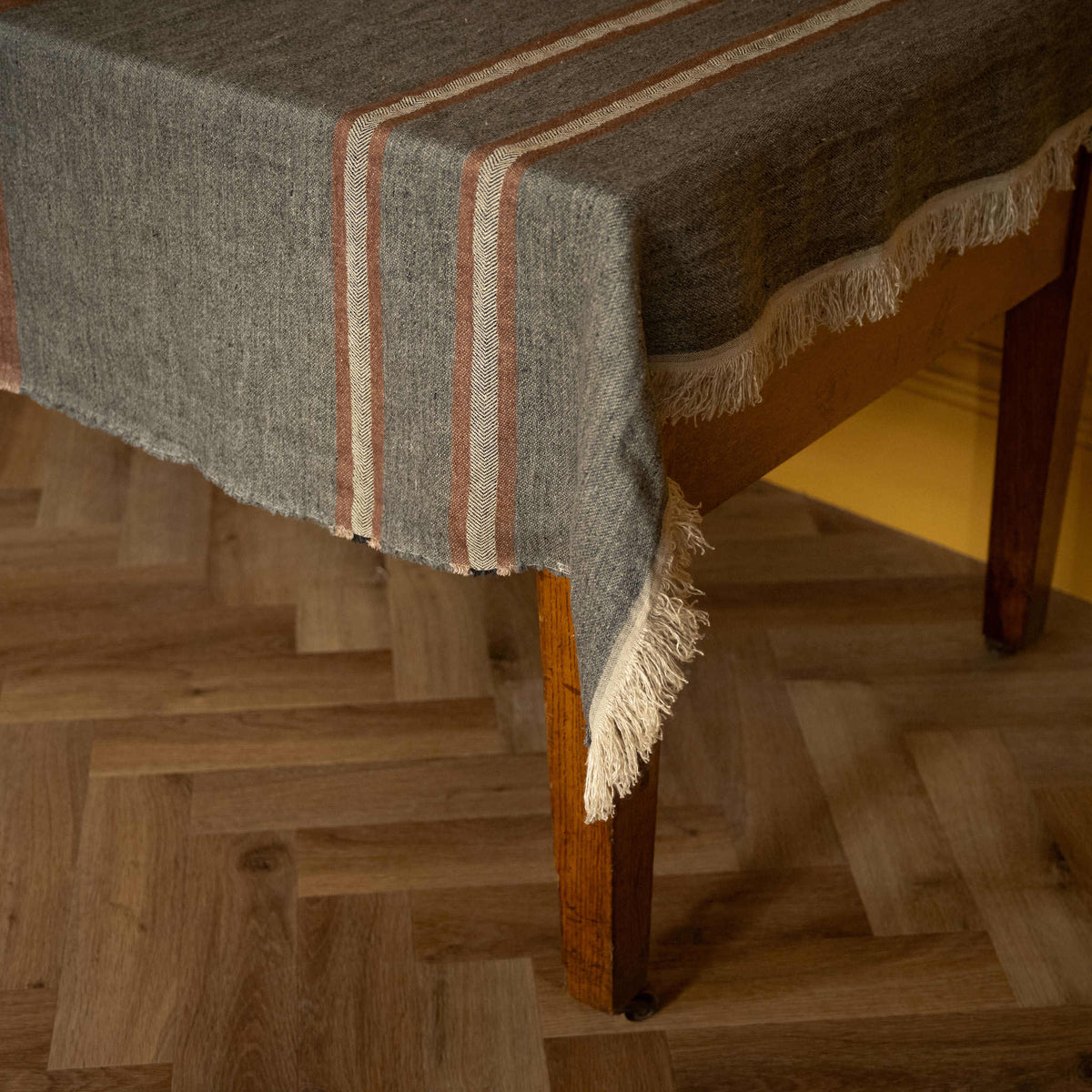 Libeco Linen Belgian Towel. A heather grey woven linen throw with ecru and sienna brown stripes and fringed edges draped over a wooden table on a herringbone wood floor.