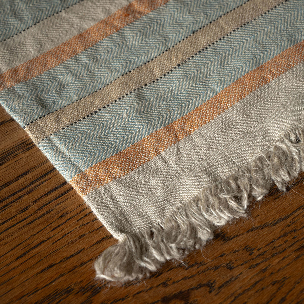 A close-up of Libeco Belgian Linen multi stripe woven hand towel with a herringbone pattern in muted blue-grey, beige, and rust orange, finished with fringed edges and shown on a wooden surface.