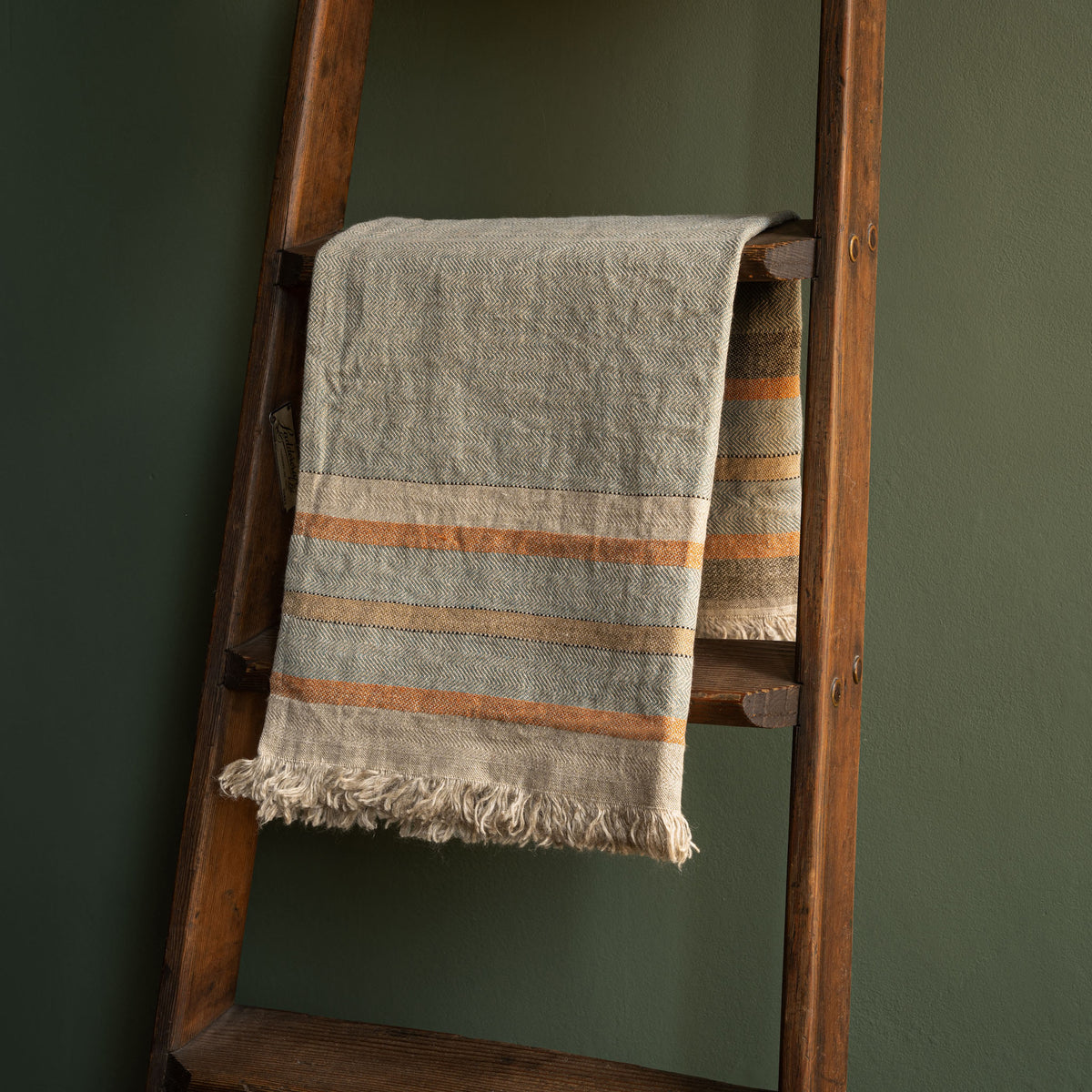 Libeco Belgian Linen multi stripe guest towel. A rustic woven hand towel in beige and muted blue-grey with orange stripes, draped over a wooden ladder against a dark green wall.