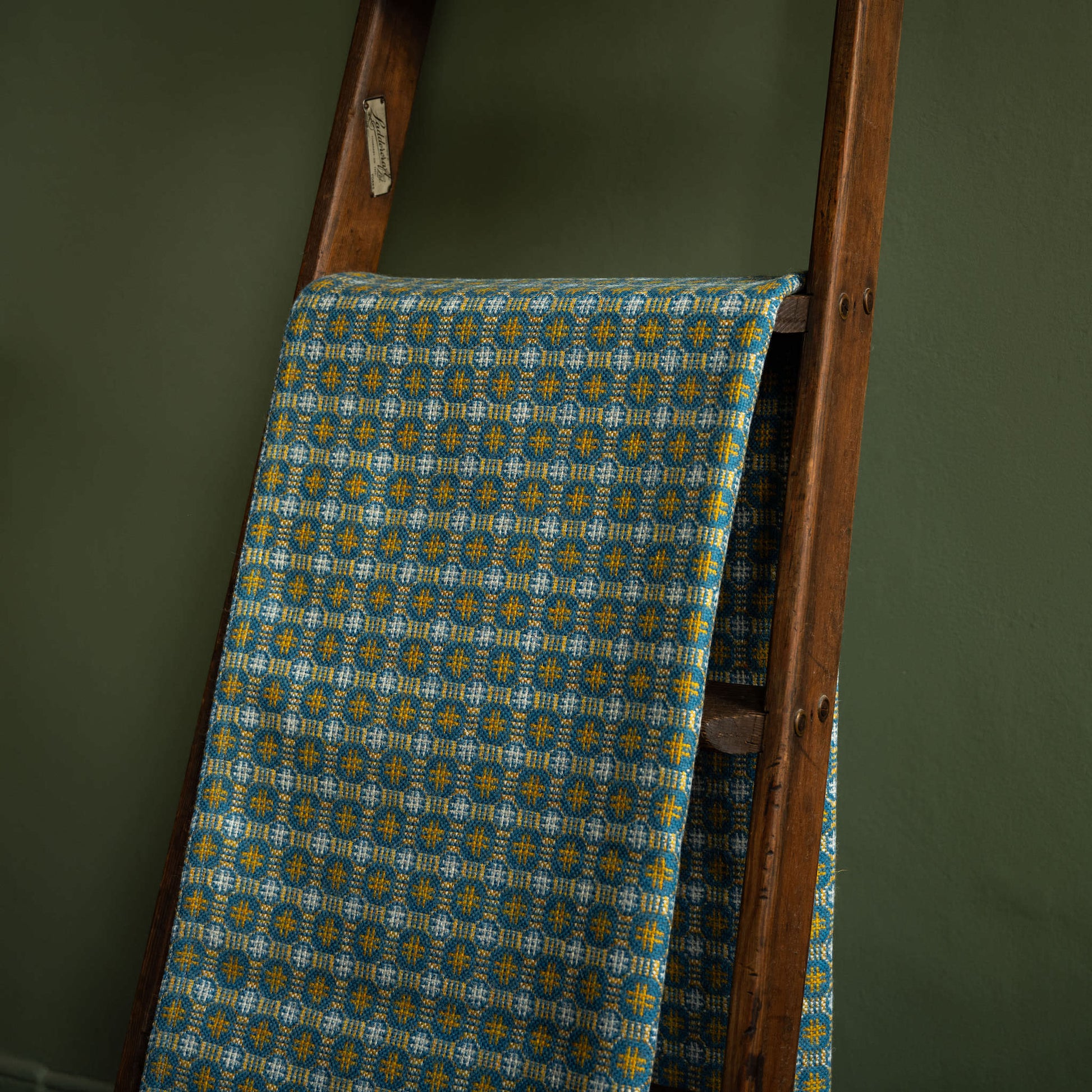 Melin Tregwynt Teils Welsh Blanket r draped over a wooden ladder against a green wall