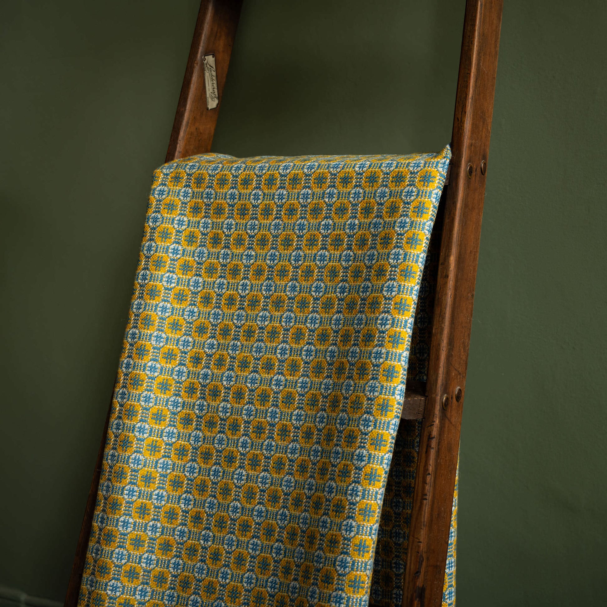 Reverse side of Melin Tregwynt Teils Welsh Blanket draped over a wooden ladder against a green wall