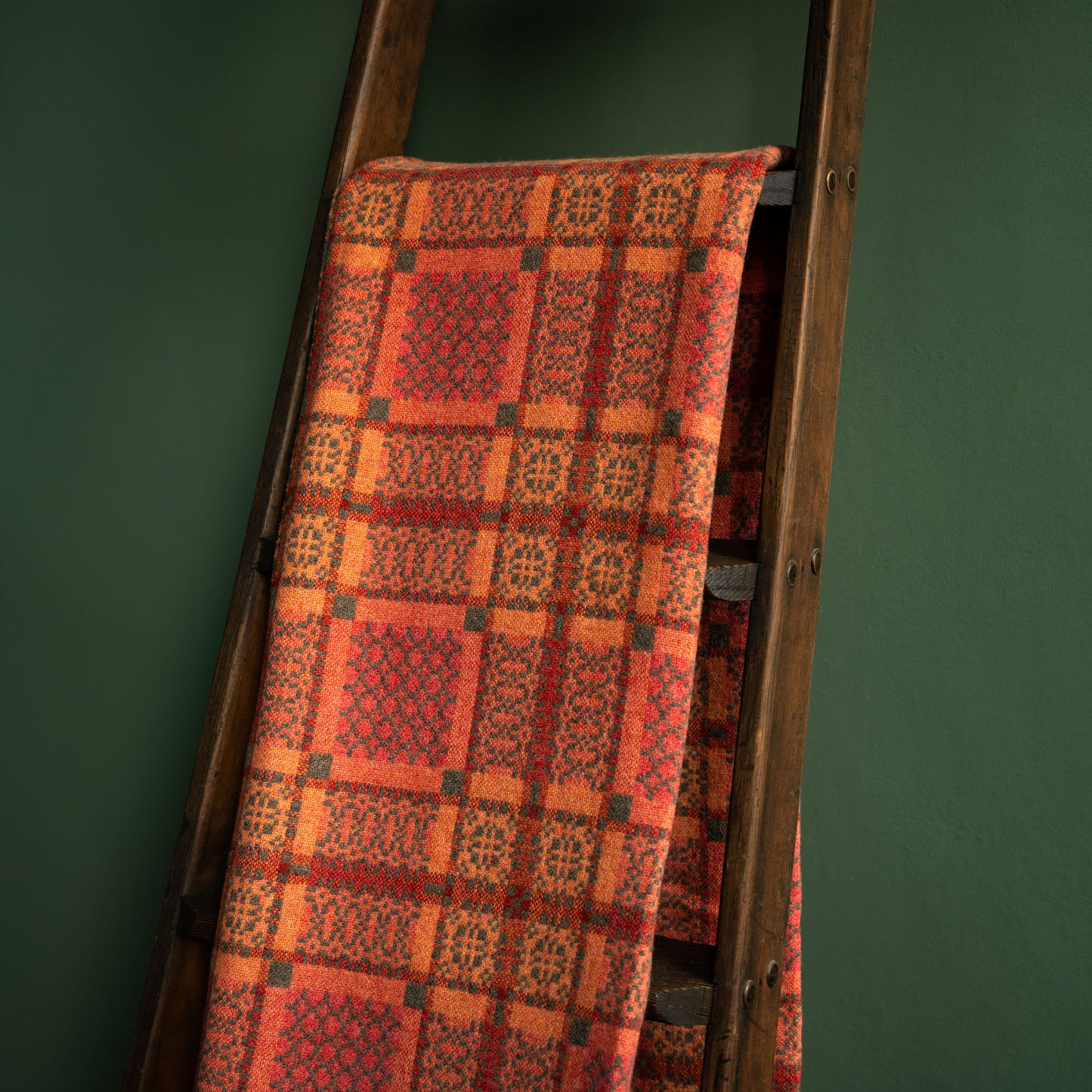 Reverse side of Melin Tregwynt copper Knot Garden Welsh wool blanket draped over a wooden ladder against a green wall