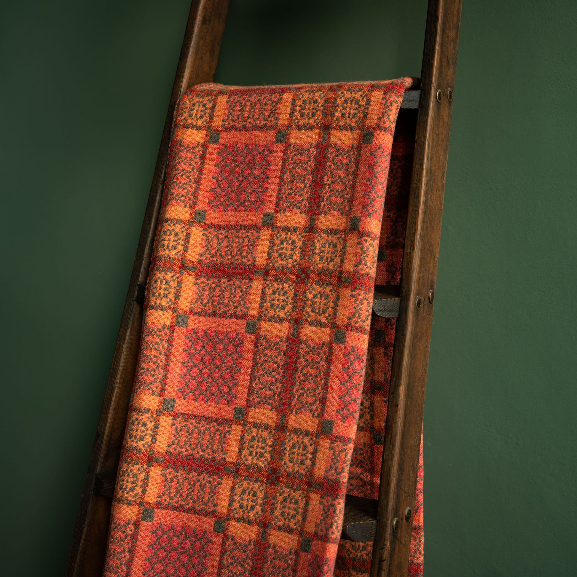 Reverse side of Melin Tregwynt copper Knot Garden Welsh wool blanket draped over a wooden ladder against a green wall
