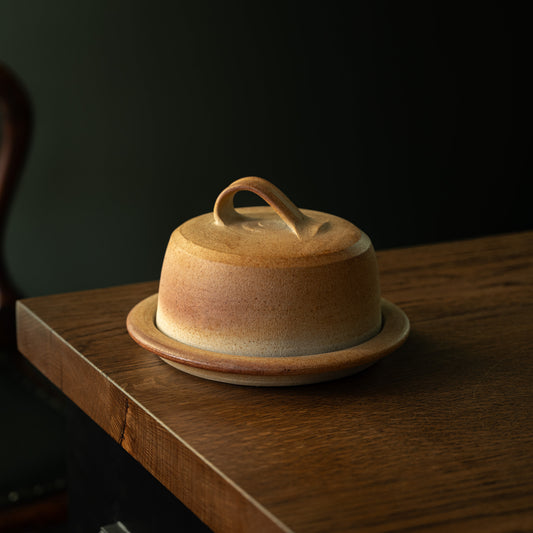 Muchelney Pottery handmade ceramic lidded butter dish with a rounded dome shape and loop handle, resting on a matching saucer in a wood-fired biscuit glaze.