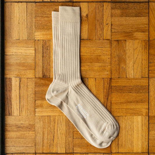 Nishiguchi Kutsushita ribbed silk and cotton sock in an oatmeal colour, laid flat on a  wooden parquet surface. The brand name 'Nishiguchi Kutsushita' is printed in white on the sole of the sock.