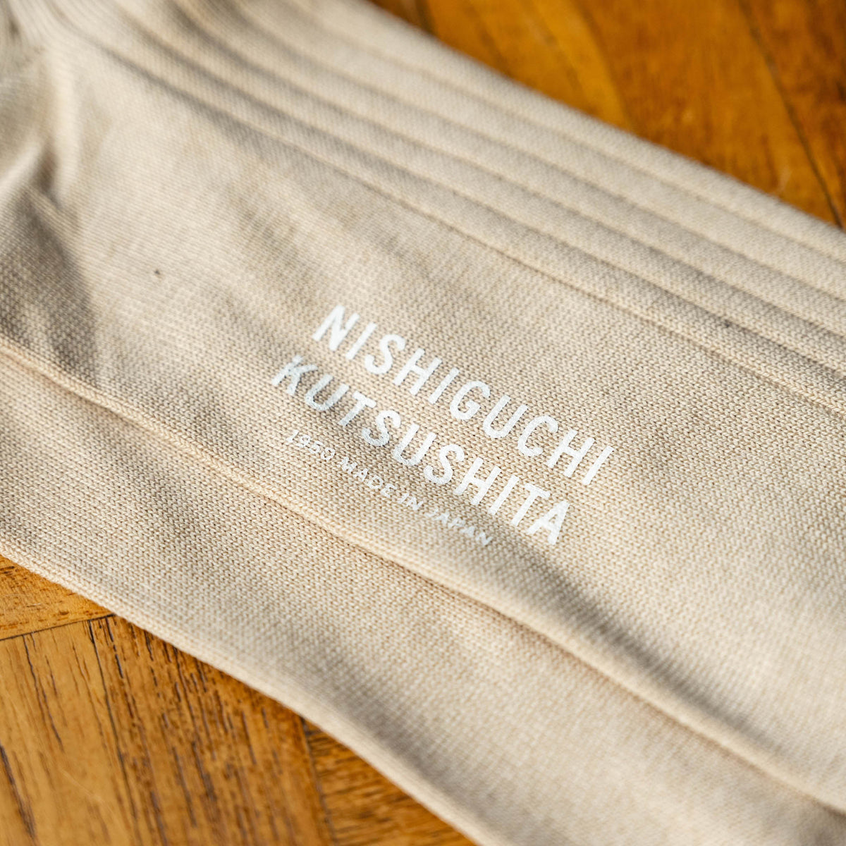 A close-up Nishiguchi Kutsushita silk and cotton ribbed socks in an oatmeal colour, showing the sole of the sock with the brand logo printed in white .