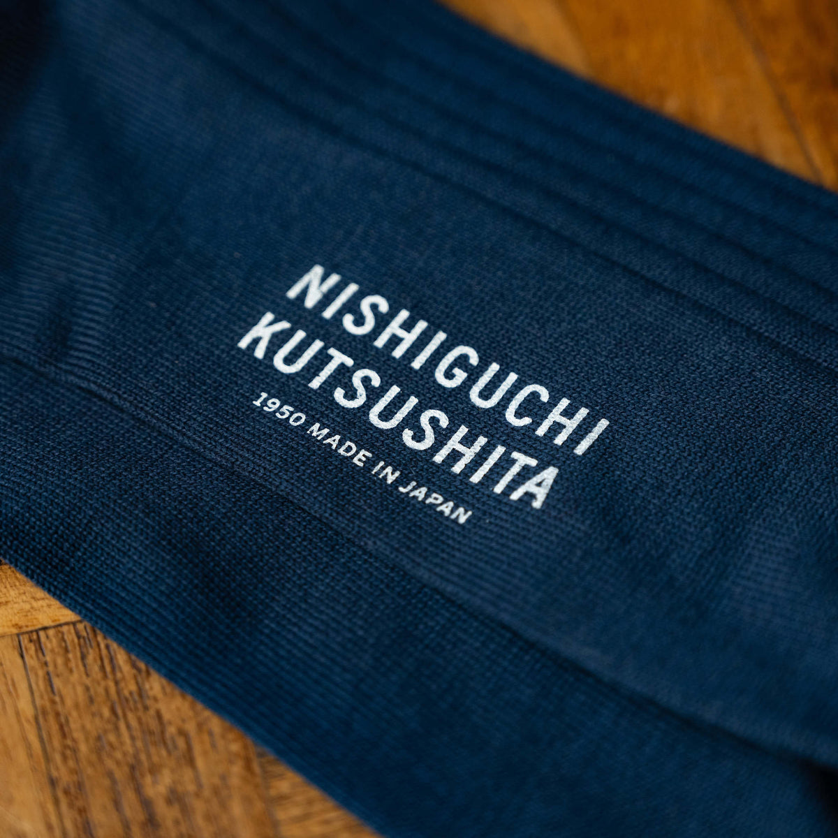 A close-up of Nishiguchi Kutsushita  dark navy blue silk and cotton ribbed socks, featuring white text on the sole that reads 'NISHIGUCHI KUTSUSHITA' '1950 MADE IN JAPAN', laid flat on a wooden surface.