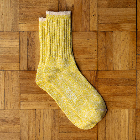 Nishiguchi Kutsushita yellow silk and cotton crew sock with a contrast colour ribbed cuff trim, laid flat on a brown wooden parquet surface.