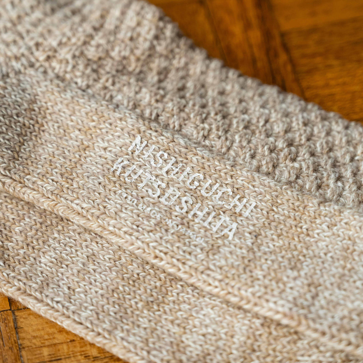 Close-up of the sole of Nishiguchi Kutsushita beige wool and cotton knit socks with white text reading 'NISHIGUCHI KUTSUSHITA'.