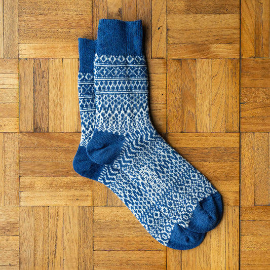 A pair of Nishiguchi Kutsushita blue and white wool jacquard patterned knit socks laid flat on a wooden parquet surface.