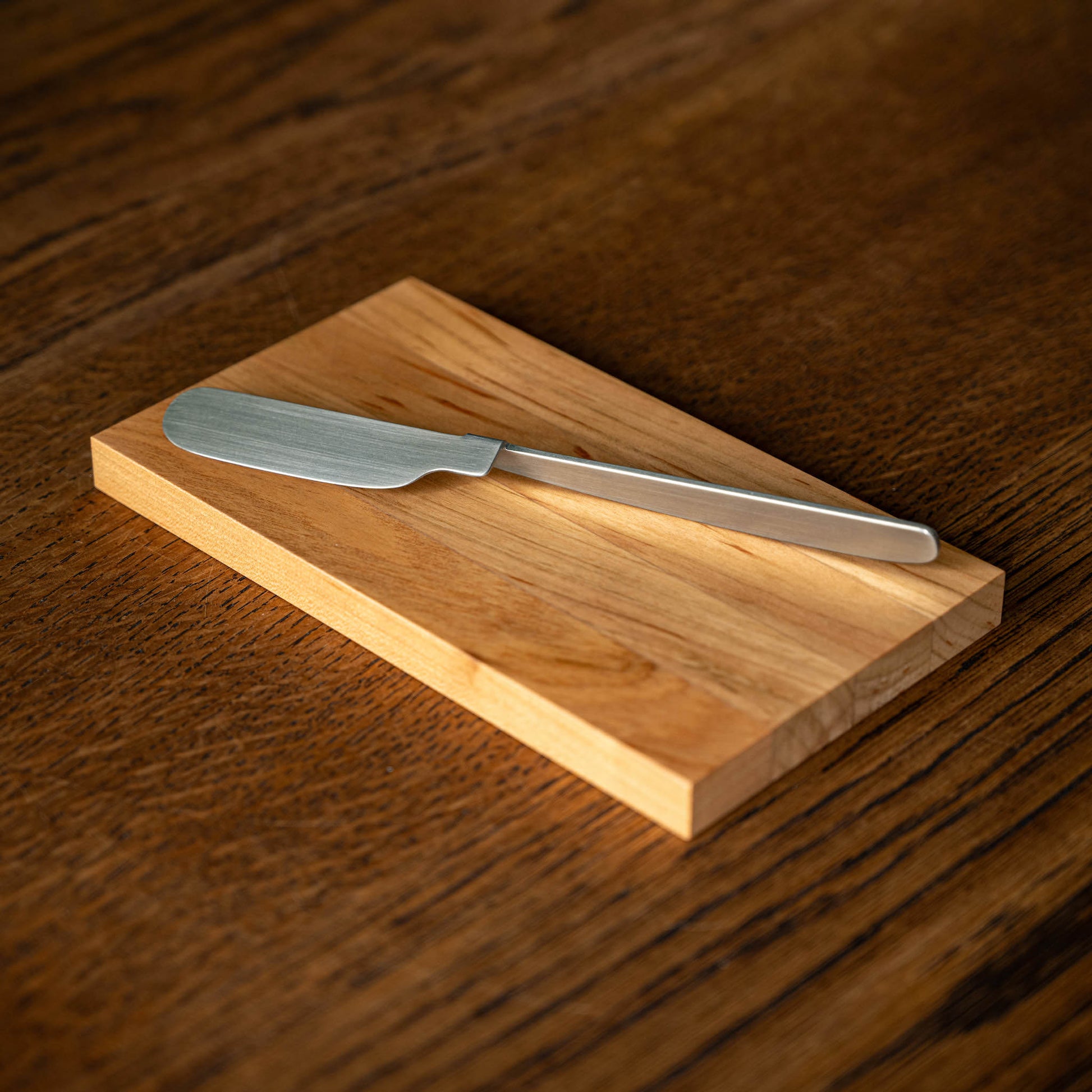 OKU Board. Designed by Kathleen Reilly. A folded stainless steel spreader knife resting diagonally on a the rectangular OKU wooden cutting board.