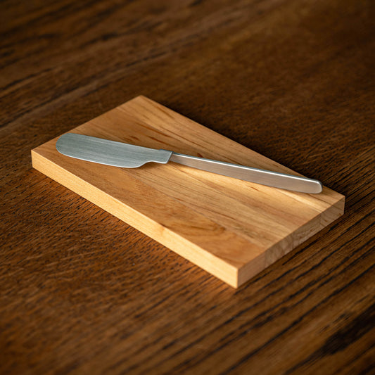 OKU Board. Designed by Kathleen Reilly. A folded stainless steel spreader knife resting diagonally on a the rectangular OKU wooden cutting board.