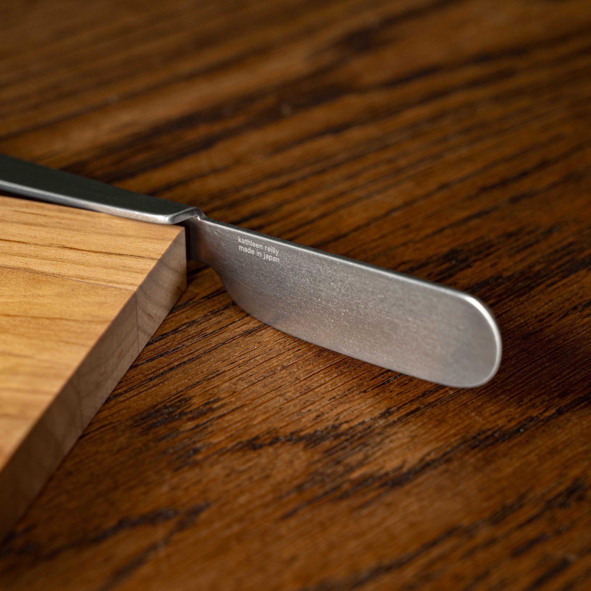 A close-up of a OKU stainless steel butter knife spreader blade resting on OKU wooden board.