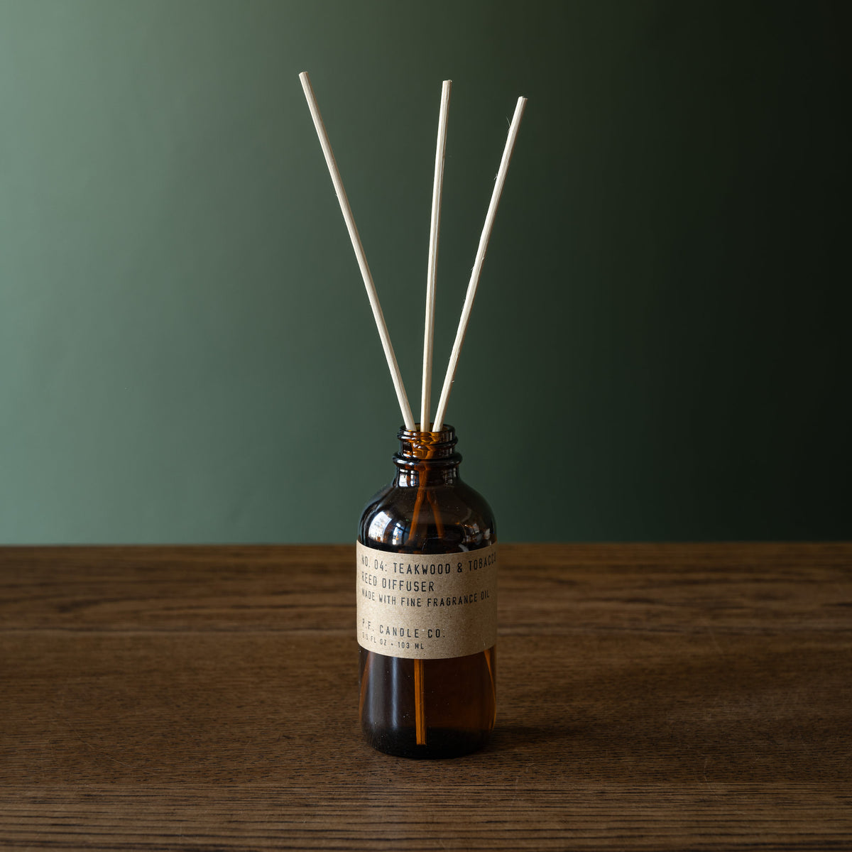 PF Candle Co Teakwood and Tobacco amber glass reed diffuser bottle with natural rattan reeds, set on a wooden surface against a dark green background. 
