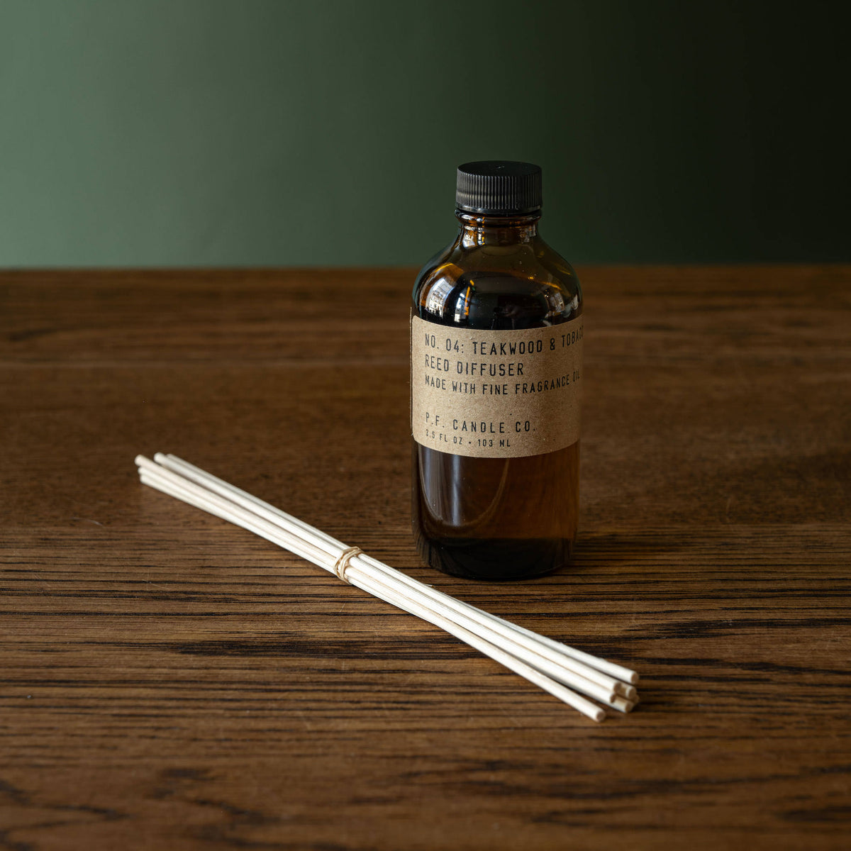 PF Candle Co Teakwood and Tobacco amber glass reed diffuser bottle with a black cap and a set of natural rattan reeds.