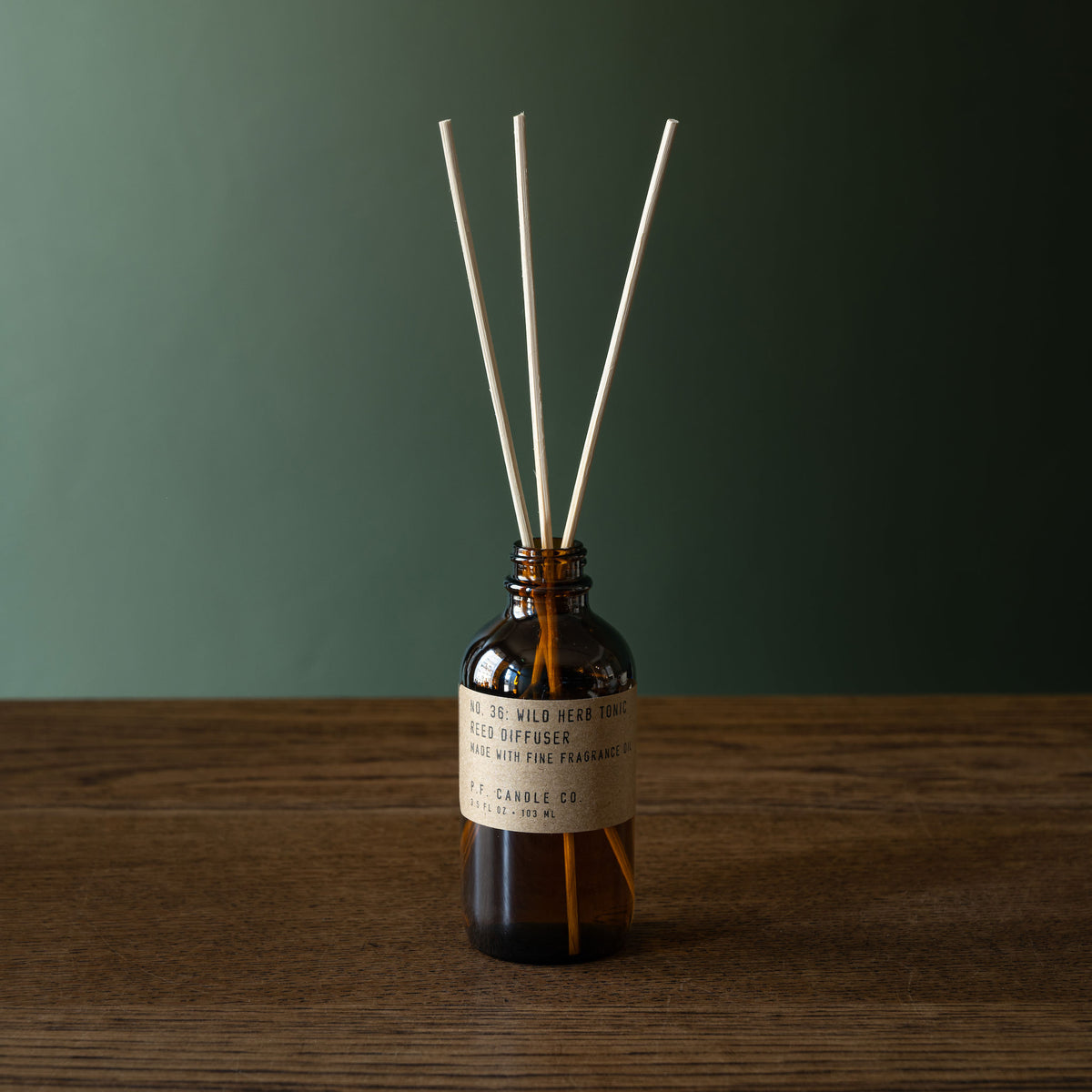 PF Candle Co Wild herb Tonic amber glass reed diffuser bottle with natural rattan reeds, set on a wooden surface against a dark green background. 