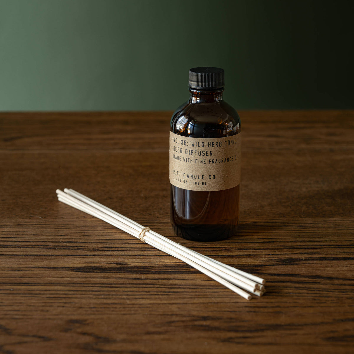 PF Candle Co Wild Herb Tonic amber glass reed diffuser bottle with a black cap and a beige brand label and set of rattan natural reeds. 