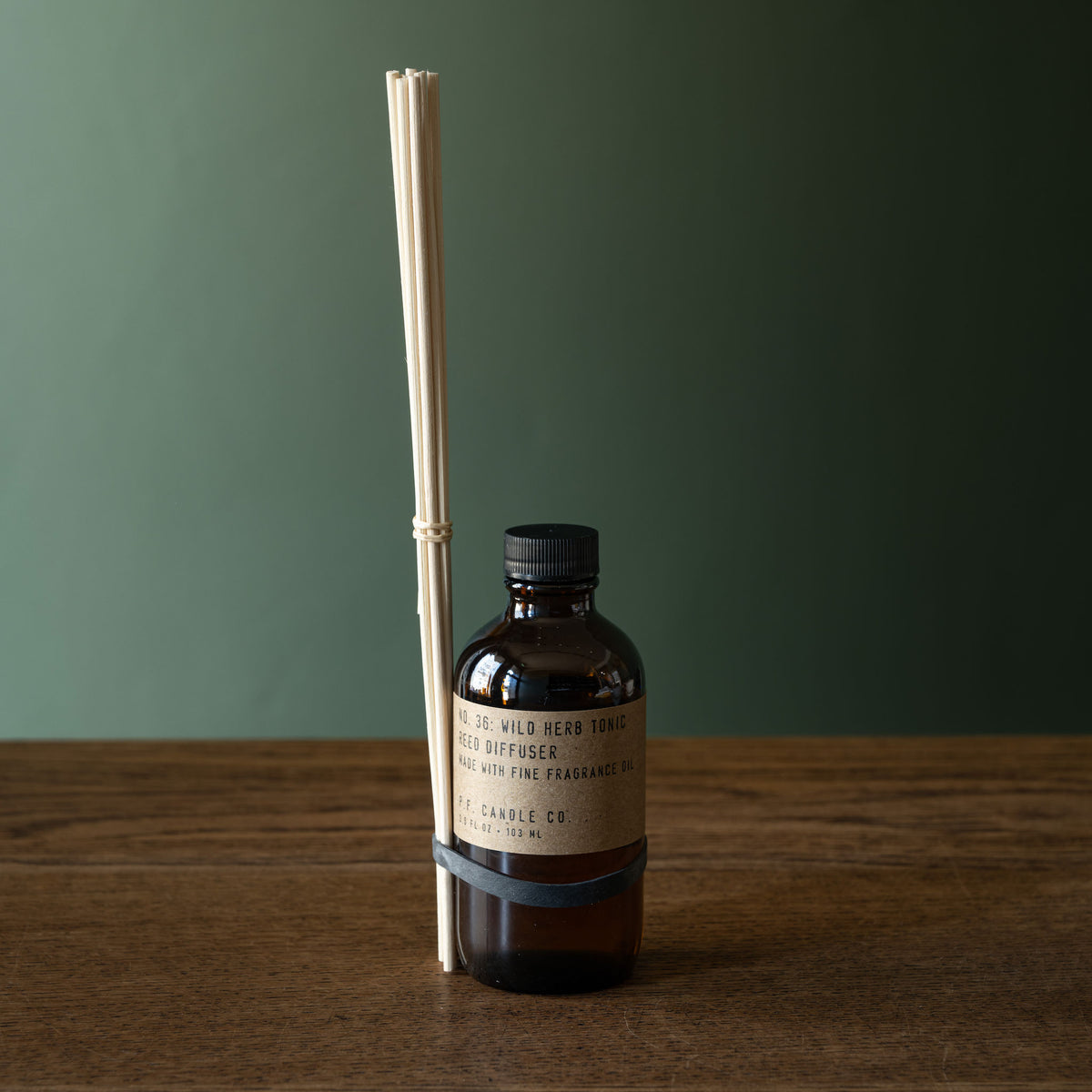 PF Candle Co Wild Herb Tonic amber glass reed diffuser bottle with a black cap and a beige label, standing on a wooden surface against a muted green background. A bundle of natural reeds is tied to the bottle with a dark band.