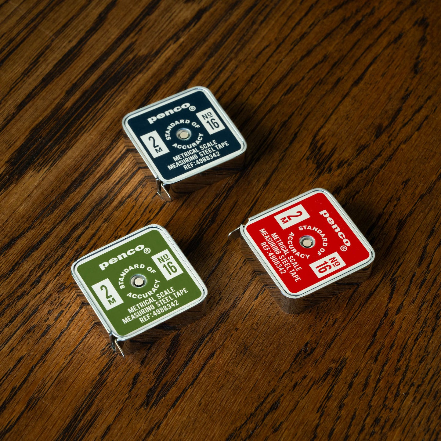 Penco pocket tape measures, navy, green and red versions on a wooden surface.