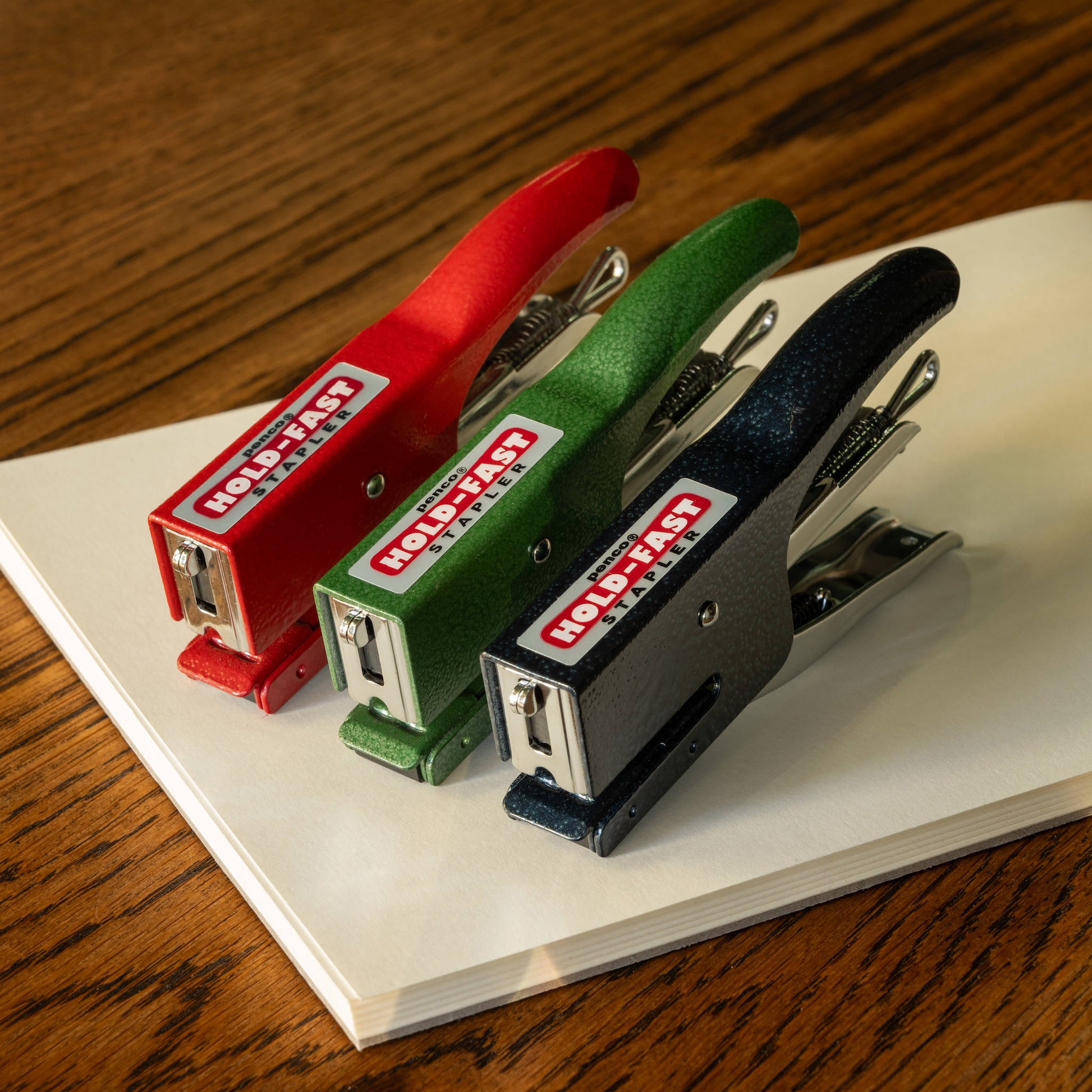 Three colourful Penco staplers on a book with a wooden surface background