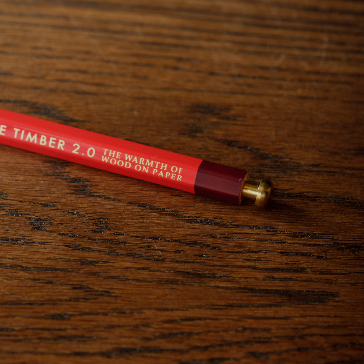 Close up of Penco Prime Timber red pencil with gold lettering reading resting diagonally on a dark brown wooden surface.