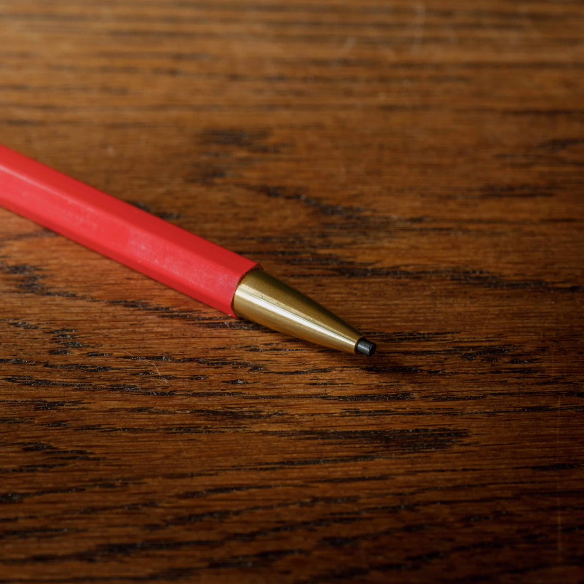 Close-up of Penco Prime Timber 2.0 red mechanical pencil lead with a brass metal tip resting on a dark brown wooden surface.