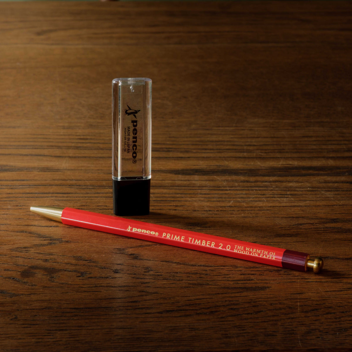 Penco Prime Timber pencil in red and a clear cylindrical sharpener case with a black lid, both branded 'Penco', placed on a dark wood desk.