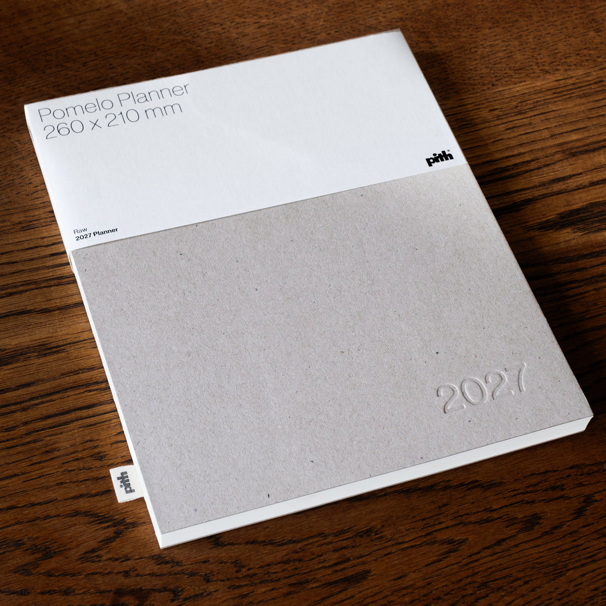 Pith Supply desk top Pomelo Planner 2027 featuring a white paper sleeve with black text and a textured raw paperboard front cover embossed with '2027', shown on a dark wood background.
