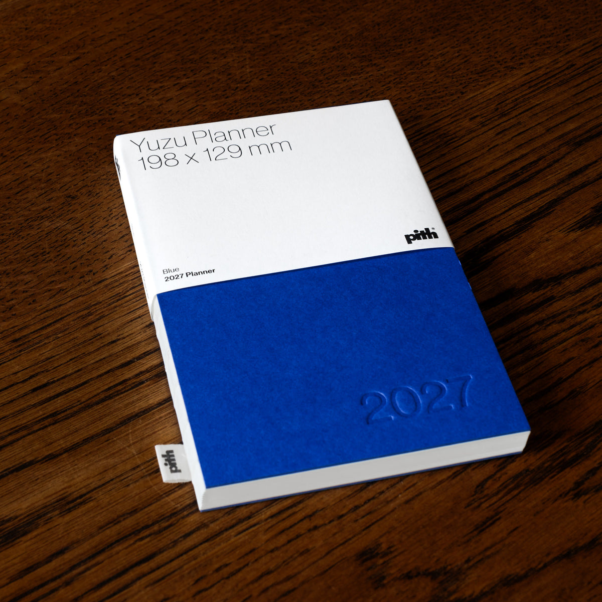 Pith Supply Yuzu Planner 2027 notebook with a blue cover, placed diagonally on a dark wood surface. 