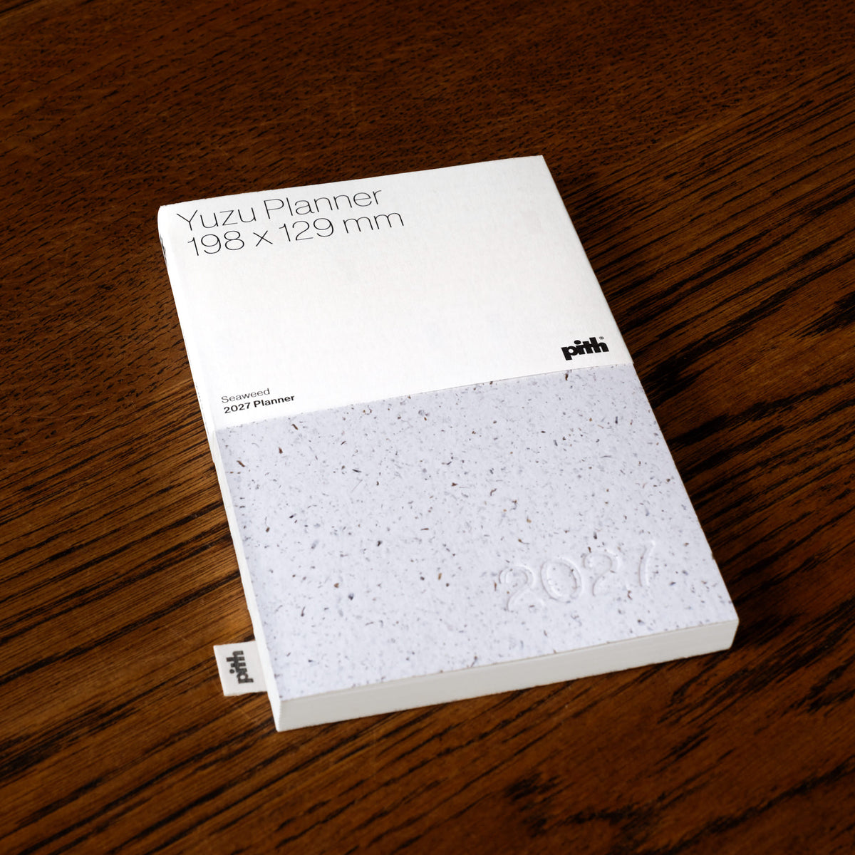 Pith Supply Yuzu Planner 2027 notebook lying on a dark brown wooden table. The cover is made from seaweed paper, with a textured feel in a speckled light grey colour. 