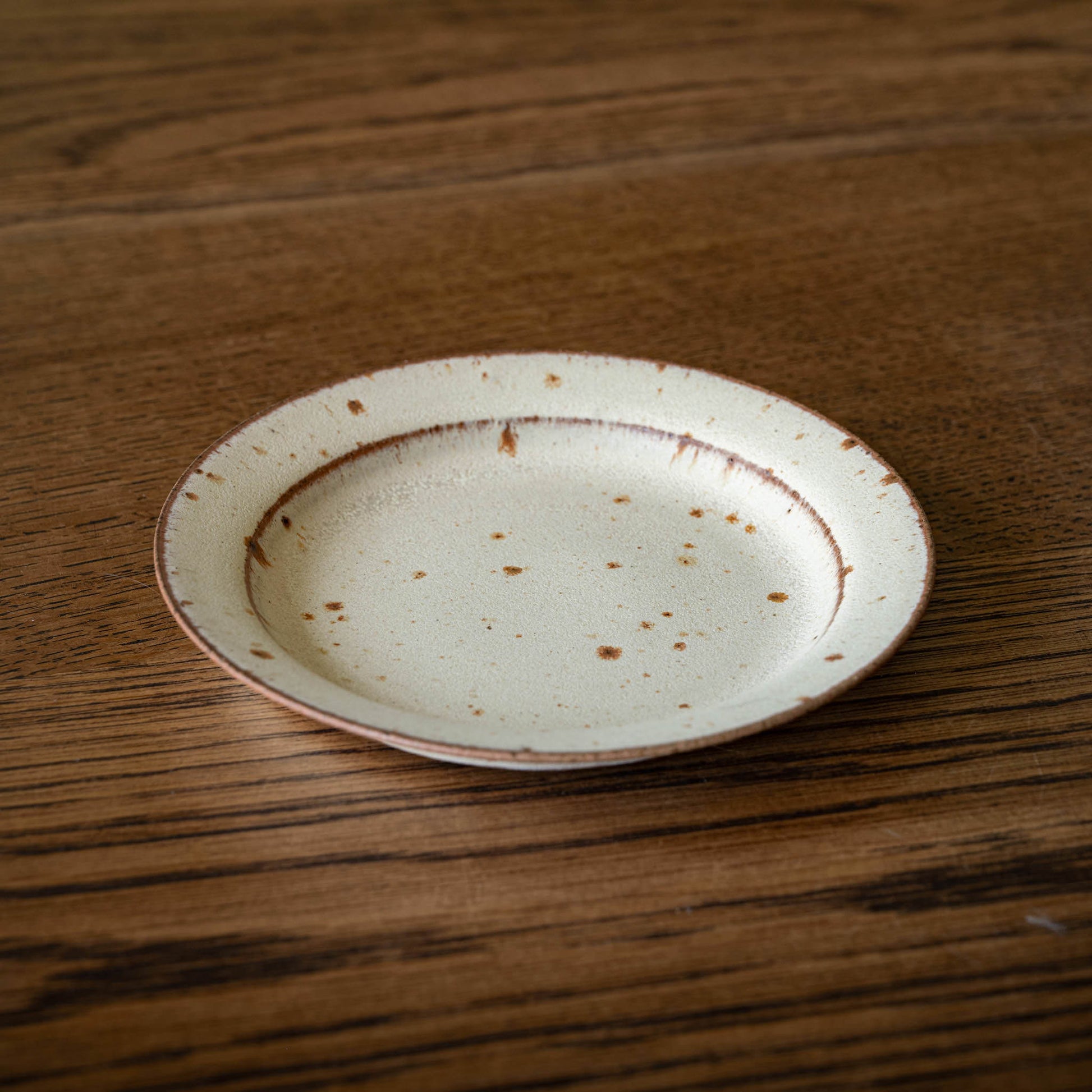 A small round ceramic biscuit plate by Studio Brae with a speckled cream glaze and brown rim, placed on a dark wood tabletop.