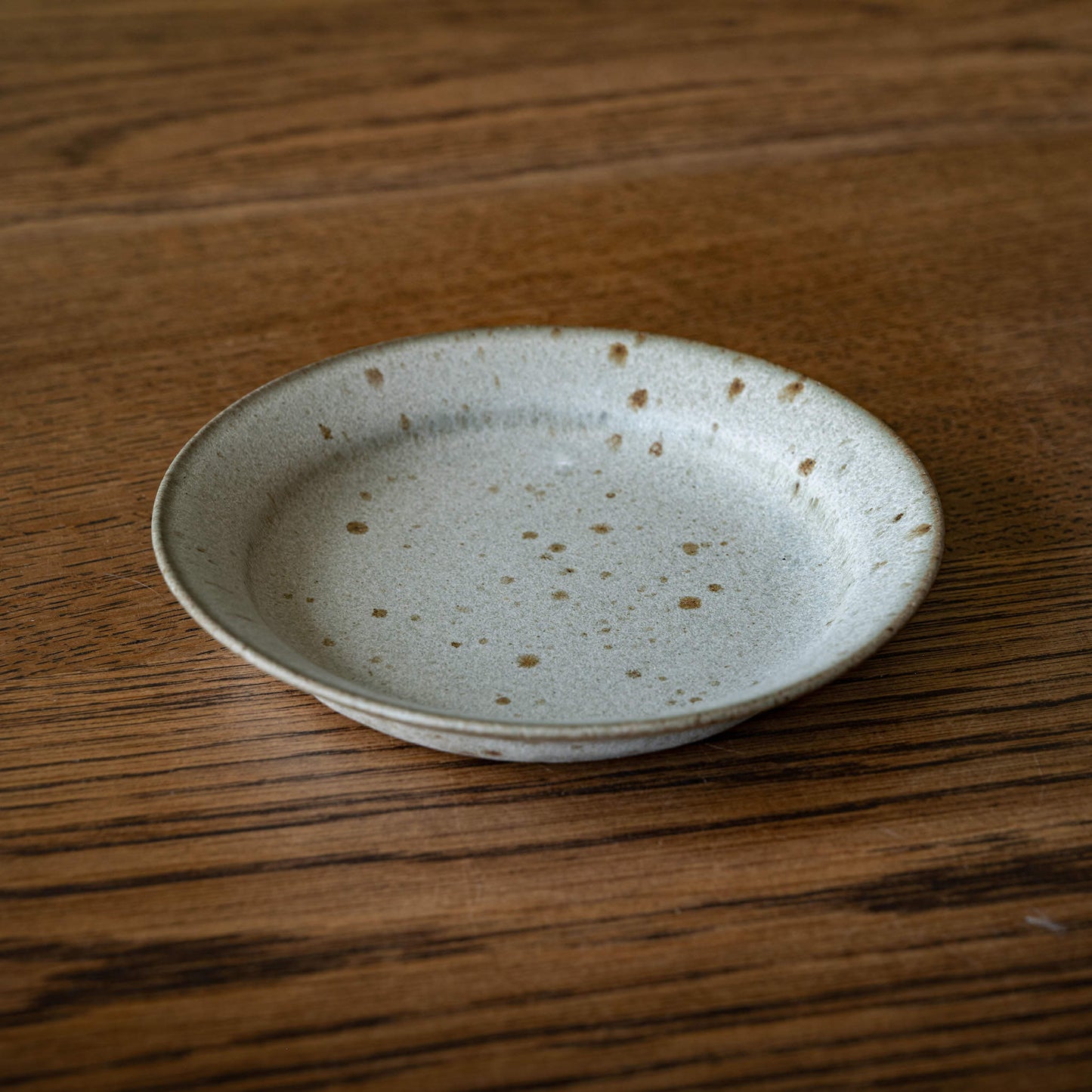 A small round ceramic biscuit plate by Studio Brae with a speckled grey and brown glaze, resting on a dark wood grain tabletop.