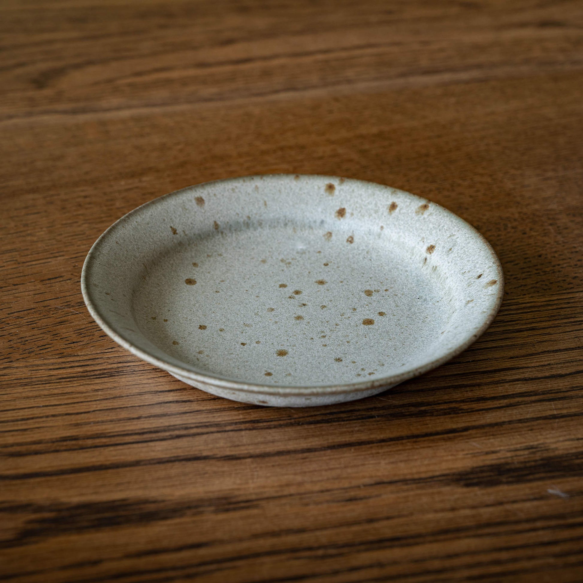 A small round ceramic biscuit plate by Studio Brae with a speckled grey and brown glaze, resting on a dark wood grain tabletop.