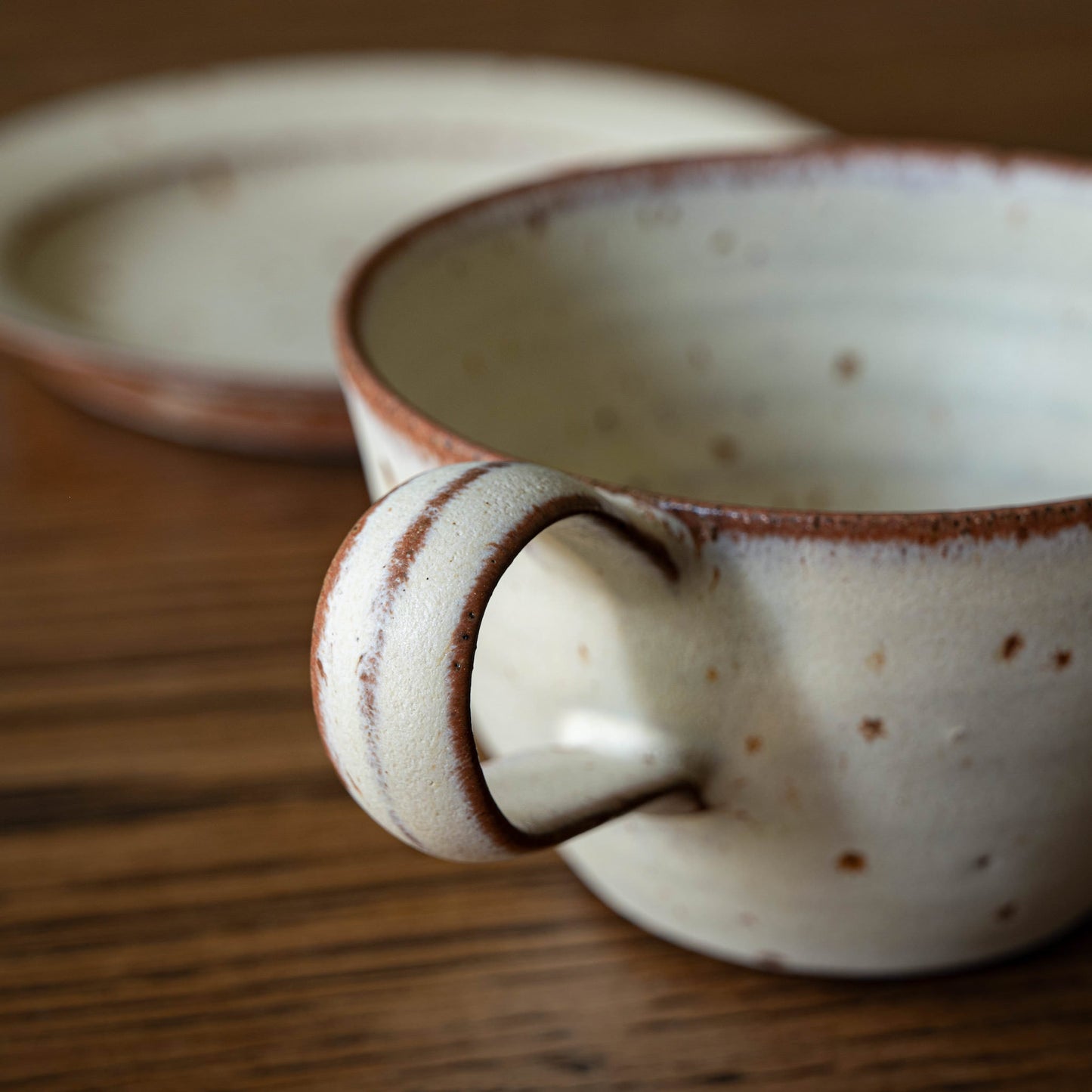 A close-up of a handmade ceramic curved cup by Studio Brae with a speckled cream glaze and brown rim, sitting on a wooden table with a matching biscuit plate in the background.