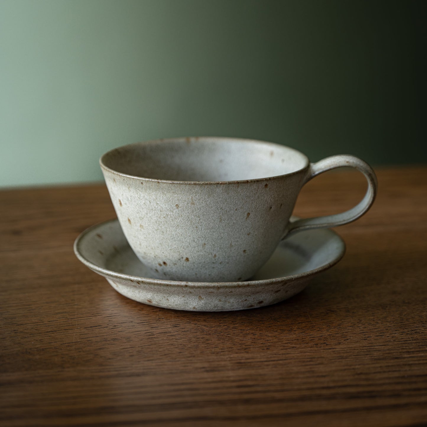 A handmade ceramic cup and biscuit plate / saucer set by Studio Brae in a speckled grey glaze, on a wooden table against a soft green background.