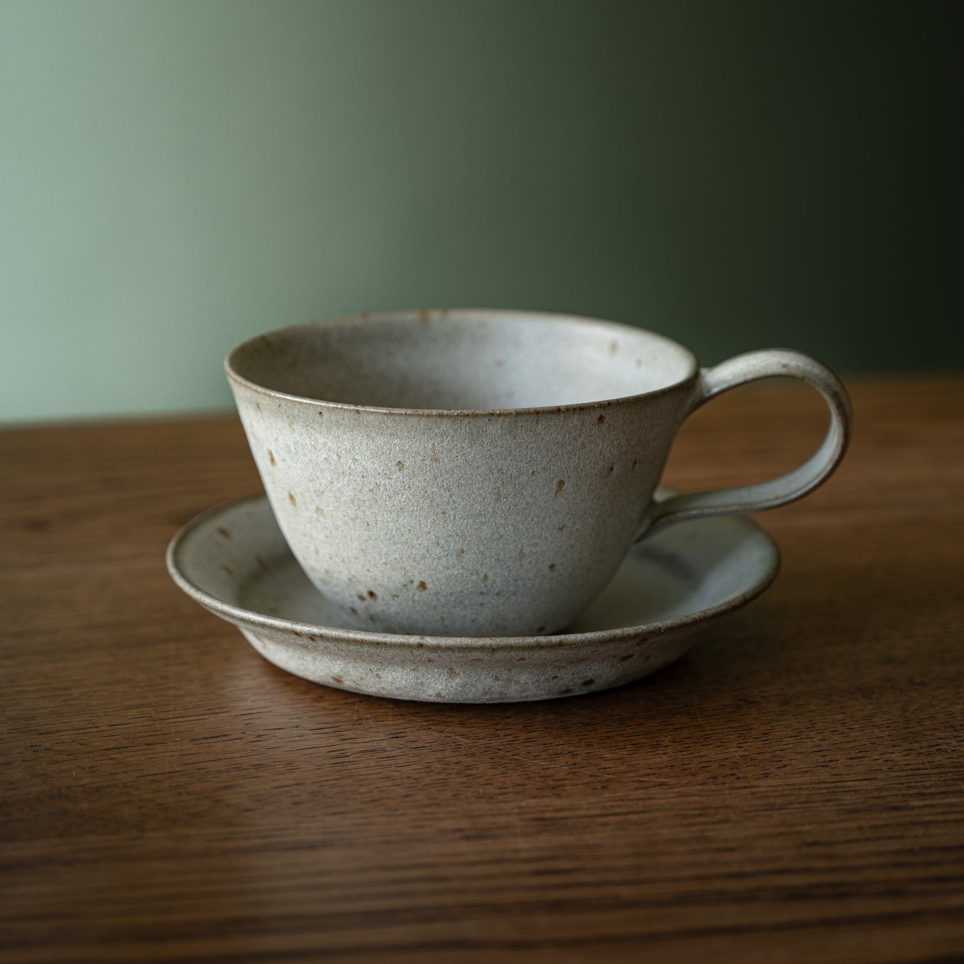 A handmade ceramic cup and biscuit plate / saucer set by Studio Brae in a speckled grey glaze, on a wooden table against a soft green background.